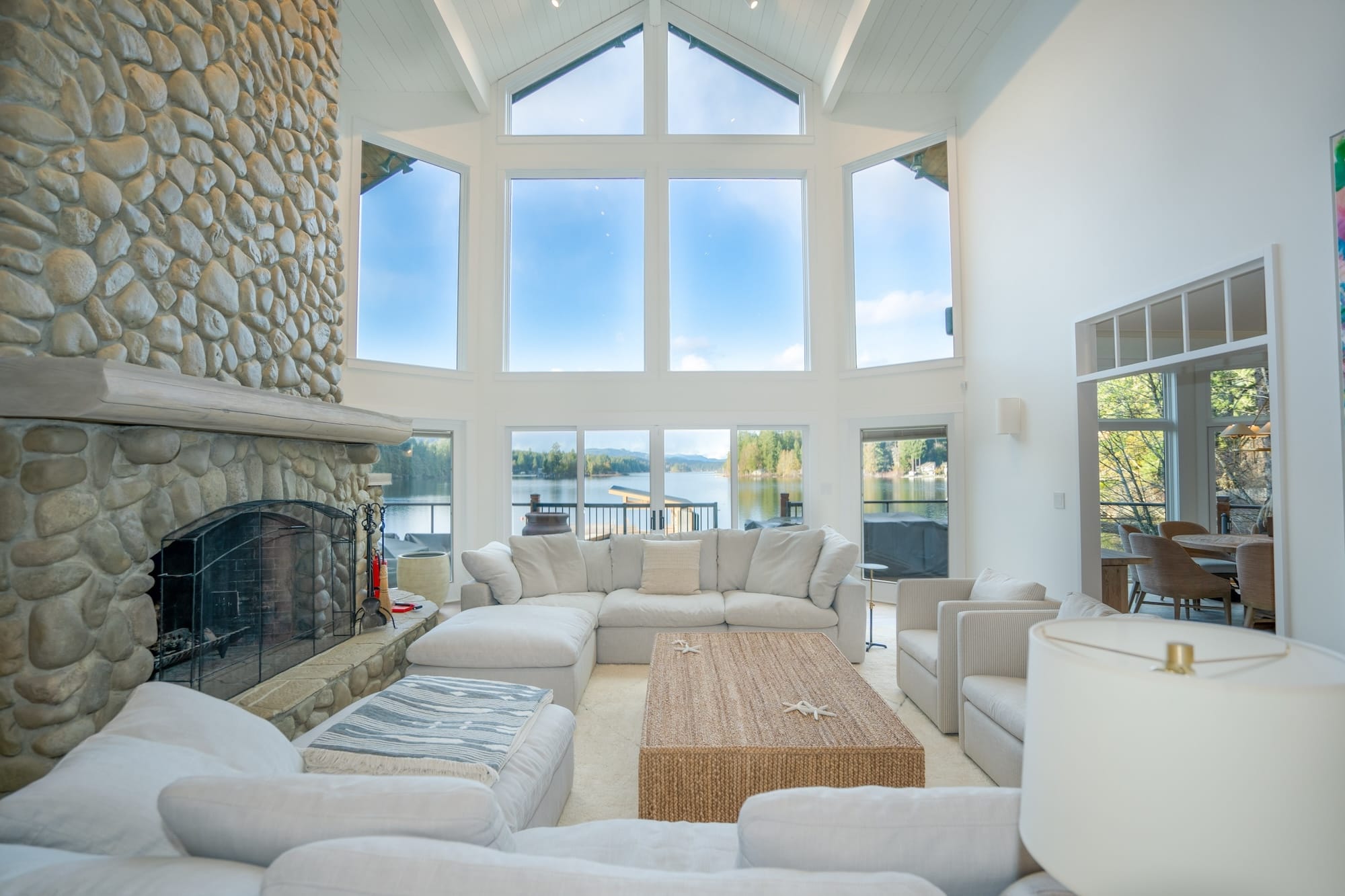 The great room — open to the rafters, lake-facing, fireplace and the dining table that runs the offsite